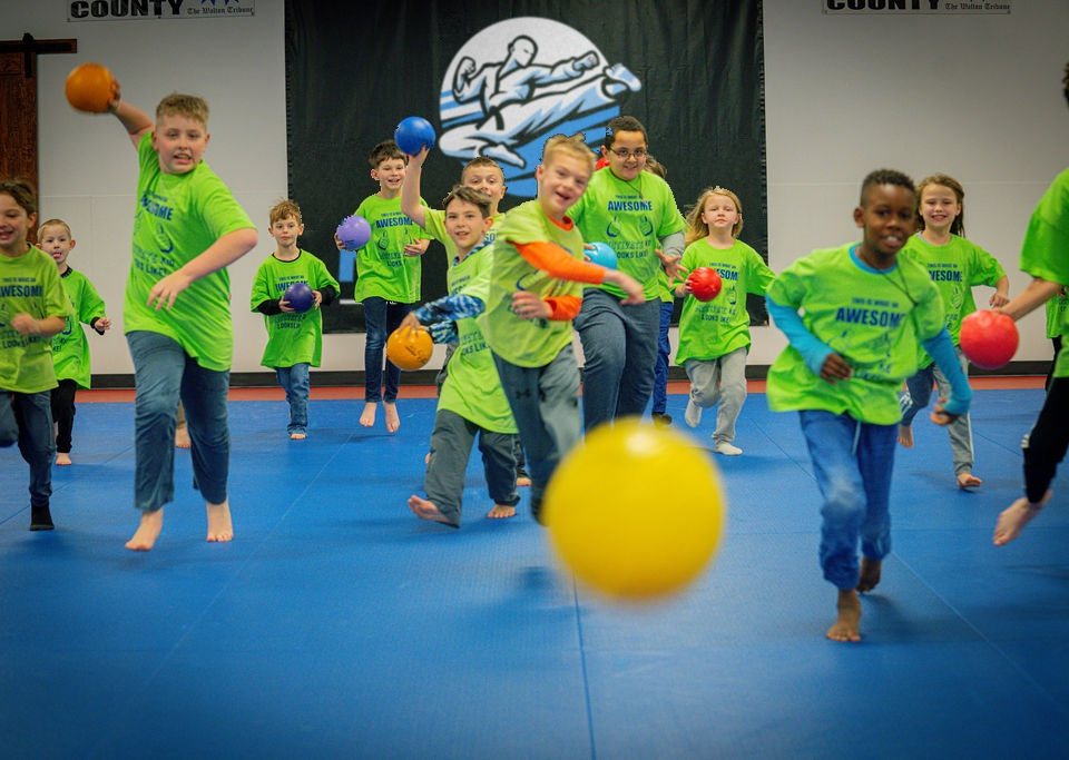 Campers in lime Motivate shirts running and throwing dodgeballs on the blue mats in front of the Motivate Martial Arts banner
