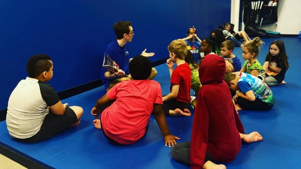 A Motivate staff member in a blue polo reading a book to a group of kids sitting on the mat